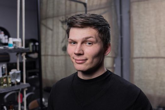 Portrait of a young positive guy of European appearance with dark hair in a black sweatshirt against the background of a hairdresser.