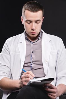 Portrait of a young intern doctor. A man sits on a chair with a notebook and pen in his hands and writes something. Making a diagnosis. Studio portrait.