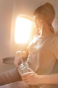 Attractive caucasian blonde girl sitting in an airplane chair looking out the window and fastens her seat belt. Safe Air Transport Concept.