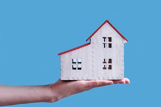 White wooden house on a female hand. Blue background. Housing concept