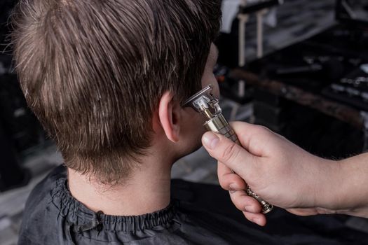 The hand of a barber or hairdresser cuts the dark long hair of the guy's client on his temple. Hairdressing.