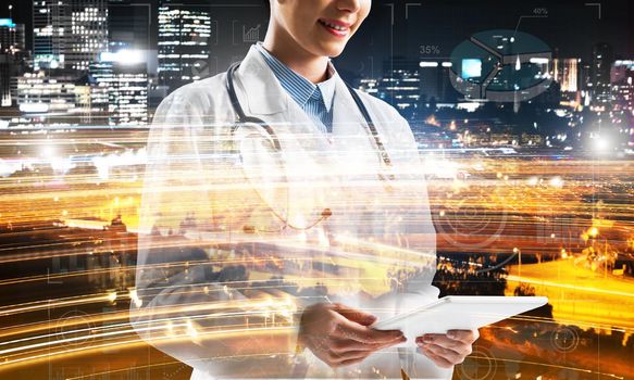 Close up of young medical industry employee standing against night city view and holding tablet in hands. Young female doctor using tablet. Double exposure with media interface icons