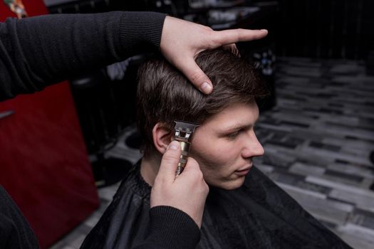 The hand of a barber or hairdresser cuts the dark long hair of the guy's client on his temple. Hairdressing.