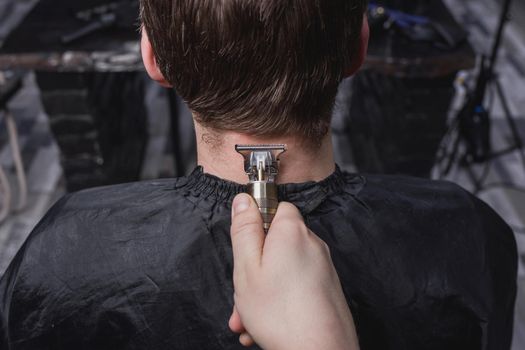 The barber's or hairdresser's hands cut the back of the head of a client's guy with dark long hair with a portable clipper. Hairdressing.