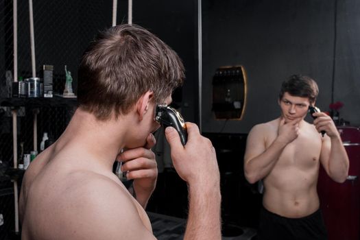 A person with a bare torso cuts his dark hair with an electric machine in front of the mirror. Care for the appearance of the house.