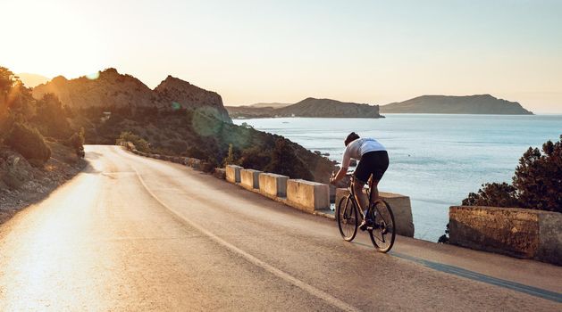 Man cyclist pedaling on a road bike outdoors in sun set at coastal road