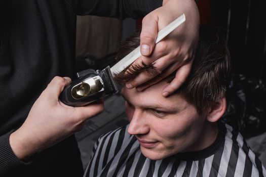 The hands of a barber or hairdresser cut a client with dark hair with an automatic machine and a comb in the barbershop. Hairdressing.