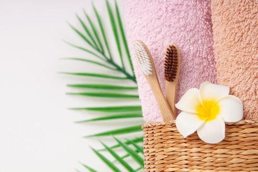 Bamboo toothbrushes with towels in a wicker wooden basket and palm leaves and plumeria flower with copy space on white background. Spa, healthy lifestyle and ecology concept.