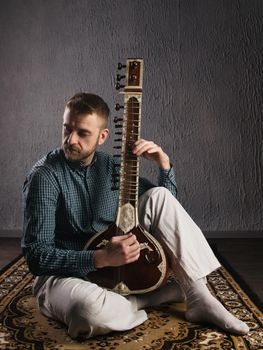 Portrait of a European man playing the sitar sitting on the carpet- Image