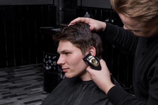 The hand of a barber or hairdresser cuts the dark long hair of the guy's client on his temple. Hairdressing.