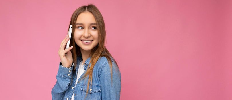 Panoramic Photo of Attractive positive smiling young blonde woman wearing casual blue jean shirt isolated over pink background holding in hand and talking on mobile phone looking to the side. Copy space
