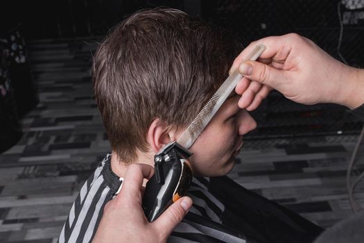 The hands of a barber or hairdresser cut the client's temple with dark hair with a automatic machine and a comb. Hairdressing.