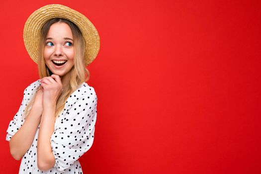 Portrait of young beautiful smiling hipster blonde woman in trendy summer dress and straw hat. Sexy carefree female person posing isolated near red wall in studio. Positive model with natural makeup. Copy space.