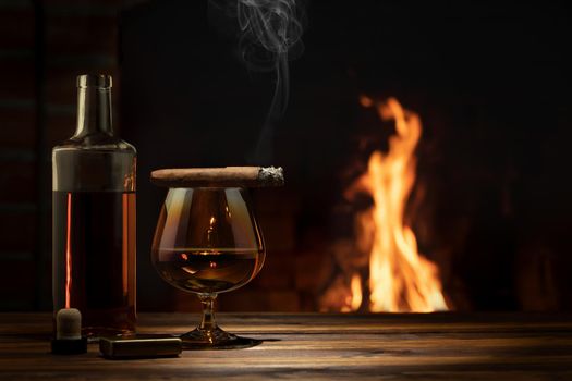 Glass of cognac, a cigar, a bottle on the table near the burning fireplace. Relaxation and enjoyment concept.