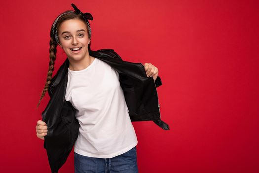 Photo of beautiful happy smiling brunette girl with pigtails wearing bandanna stylish black leather jacket and white t-shirt for mockup isolated over red background wall looking at camera. Empty space