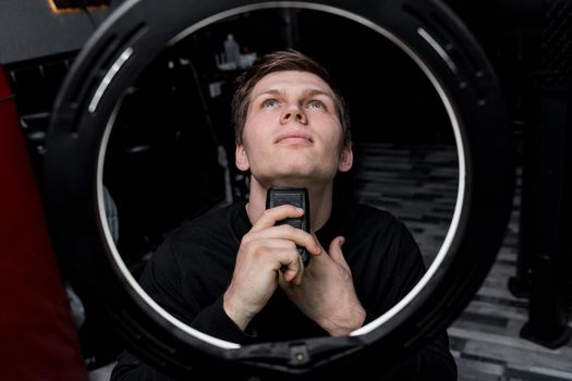 A serious young guy of Caucasian appearance with dark hair shaves with an automatic razor next to the ring lighting. Care for your appearance.