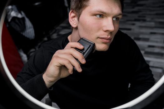 A serious young guy of Caucasian appearance with dark hair shaves with an automatic razor next to the ring lighting. Care for your appearance.