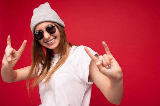Closeup photo shot of beautiful happy young dark blonde woman isolated over red background wearing casual white t-shirt for mockup and stylish sunglasses looking at camera and showing rock and roll sign.