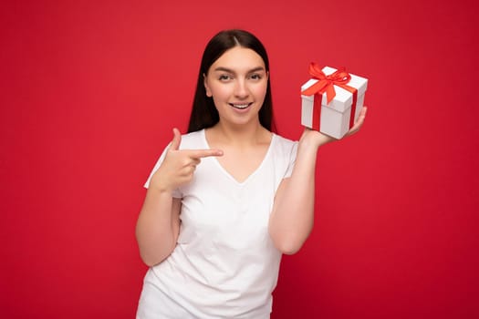 Photo of beautiful positive smiling young brunette woman isolated over red background wall wearing white casual t-shirt for mockup holding white gift box with red ribbon and looking at camera and pointing finger at present.