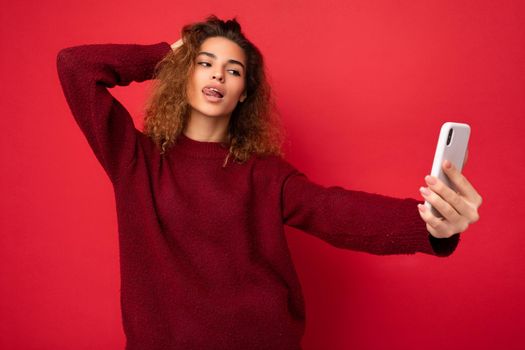 Sexy Beautiful young woman with curly hair wearing dark red sweater isolated on red background wall holding and using smart phone looking at telephone screen and taking selfie and showing tongue.