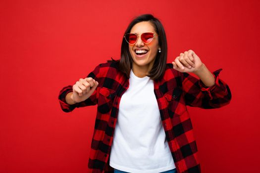 Portrait of young happy positive beautiful brunette woman with sincere emotions wearing white t-shirt, stylish red check shirt and red sunglasses isolated on red background with copy space and dancing.