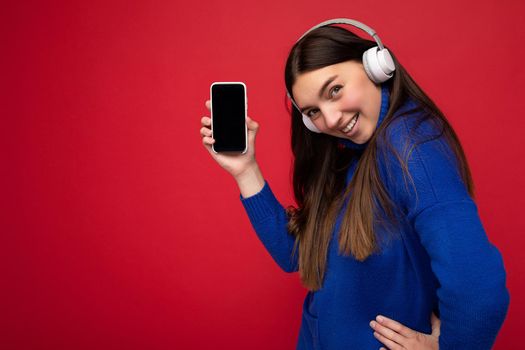 Side-profile portrait photo shot of charming happy young brunette woman wearing casual blue sweater and white earphones isolated over red background with empty space holding in hand mobile phone and showing smartphone with empty screen for mockup listening to music looking at camera.