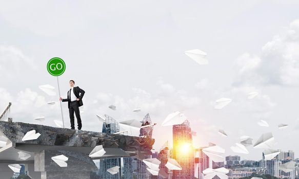 Confident businessman in suit holding green go sign while standing among flying paper planes on broken bridge with cityscape and sunlight on background. 3D rendering.