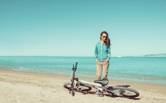 Beautiful young woman in sunglasses standing near a bicycle on sand beach in summer