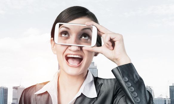Portrait of pretty woman covering her eyes with smartphone. Happy businesswoman showing mobile phone with eyes on screen. Corporate businessperson on cityscape background. Mobile communication layout.