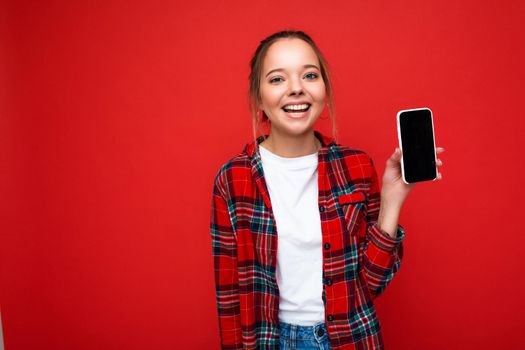 Photo of beautiful smiling young woman good looking wearing casual stylish outfit standing isolated on background with copy space holding smartphone showing phone in hand with empty screen display for mockup looking at camera.