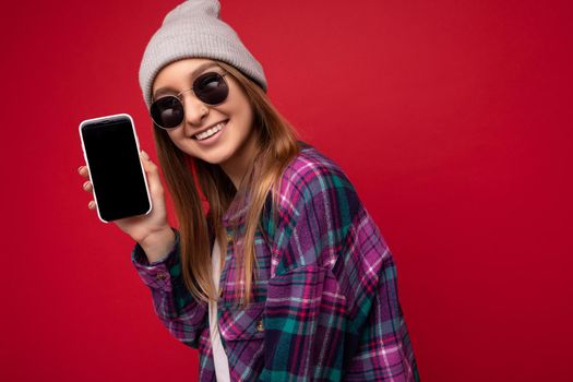 Photo of beautiful smiling young woman good looking wearing casual stylish outfit standing isolated on background with copy space holding smartphone showing phone in hand with empty screen display for mockup looking at camera.