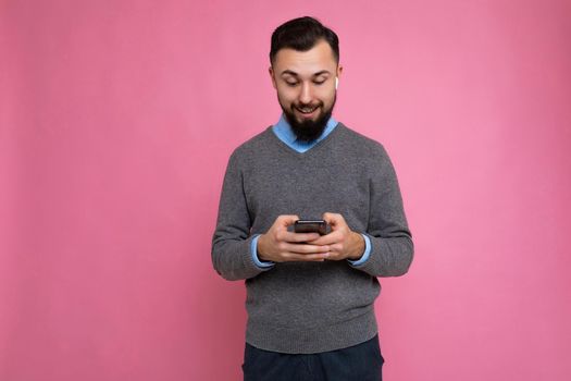 Photo shot of surprised cool good looking brunet unshaven young man with beard wearing grey sweater and blue shirt isolated over pink background wall with empty space holding in hand and using mobile phone messaging sms online looking down at device display.