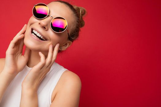 Photo shot of beautiful young smiling blonde woman wearing casual clothes and stylish sunglasses isolated over colorful background looking at camera.