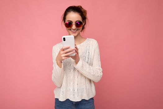 Photo shot of attractive positive good looking young woman wearing casual stylish outfit poising isolated on background with empty space holding in hand and using mobile phone messaging sms looking at smartphone display screen.