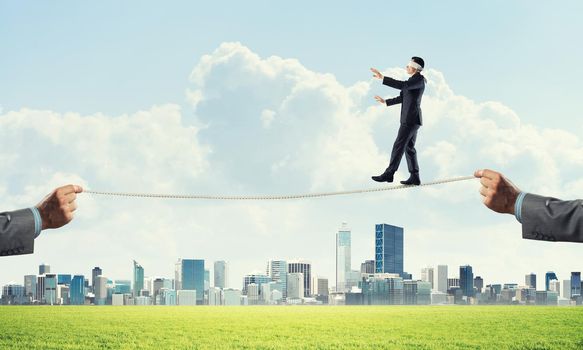 Businessman with blindfolder on eyes walking on rope over cityscape background