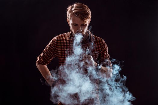 young man wearing a plaid shirt smokes an electronic cigarette on a black background