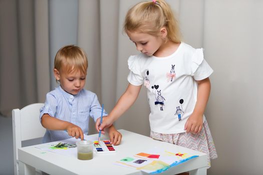 The boy and girl, brother and sister paint watercolors at the table on white sheets of paper. Concept aesthetic development of children, happy childhood.