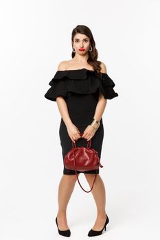 Beauty and fashion concept. Full length if silly young woman pouting and looking confused, holding purse, wearing heels and black dress, white background.