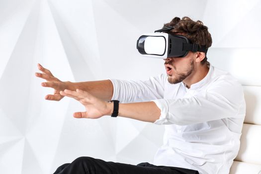 Excited young man is getting experience using VR-headset glasses of virtual reality gesticulating with his hands. A young man sits on a comfortable armchair in a room with white walls