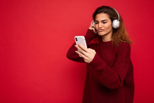 Photo shot of beautiful dissatisfied young female person wearing stylish casual outfit isolated over colorful background wall wearing white bluetooth wireless earphones and listening to music and using mobile phone looking at gadjet display. copy space