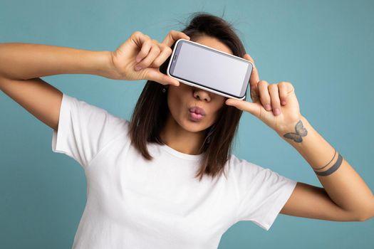 Photo of pretty positive young brunette woman good looking wearing white t-shirt standing isolated on blue background with copy space holding smartphone showing phone in hand with empty screen screen for mockup and giving kiss.