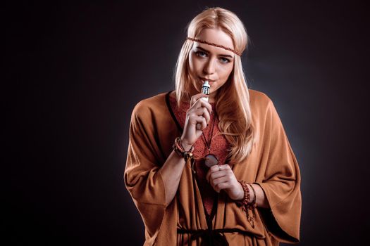 Young woman in the Boho style blowing smoke. The blonde on a dark background. Young woman looking into the camera