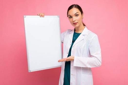 Portrait of attractive pretty young positive brunette woman wearing medical white coat holding white magnetic board for mock up isolated over pink background with copy space.