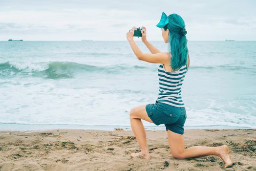 Beautiful young woman with blue hair taking photographs of sea in summer