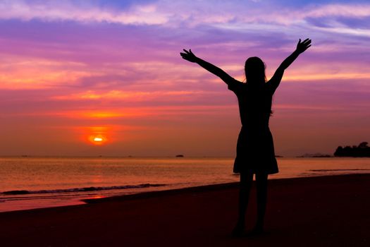 Silhouette of woman with hands up while standing on the sea beach at sunset