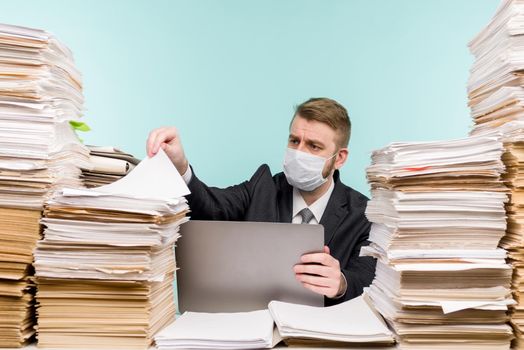 A male accountant or company manager works in an office in a pandemic in view of the accumulated paper work. A protective medical mask is on the face. On the desktop are large stacks of documents. - image