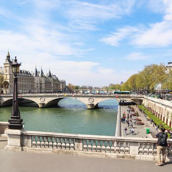 Paris / France - April 06 2019: Cityscape of Paris and Saint-Michel bridge across Seine river. A tourist photographs a beautiful view of the promenade with people resting on sunny day