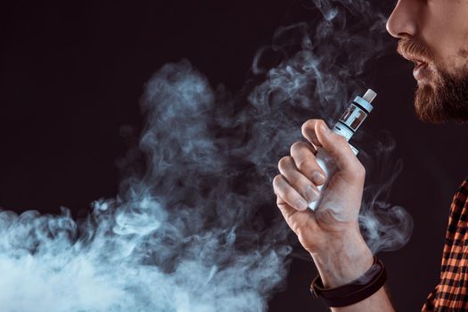 young man wearing a plaid shirt smokes an electronic cigarette on a black background. close-up