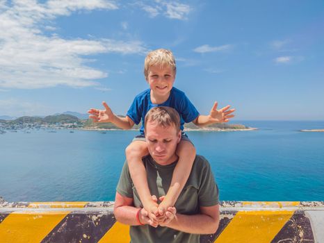 Front boy son laugh sitting on fathers shoulders. No photoshop sun on skin. Sea, clouds, island background. Funny photo, happiness lifestyle, Father's Day, love parenthood, family holiday concept.