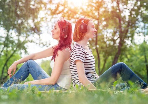 Two young women listen to music in the park. Sitting back to back